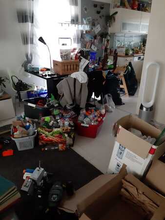 Unaufgeräumtes Wohnzimmer mit Kartons, Haushaltswaren und Verpackungen vor moderner Küche.