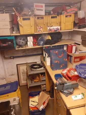 Unordentlicher Kellerraum mit Werkzeug, Kartons, gelben Kisten und Ablagen auf Regalen.